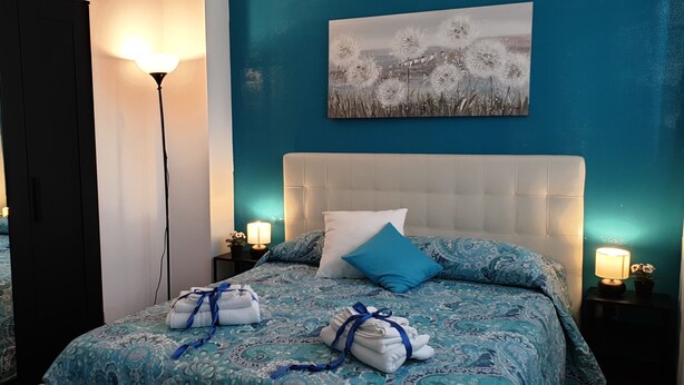 Une chambre avec un mur bleu, une tête de lit blanche et un tableau représentant des pissenlits. On y trouve deux tables de chevet avec des lampes, ainsi qu'un lit avec une literie bleue et deux serviettes.
