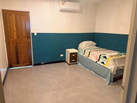 Quarto com uma cama de solteiro, porta de madeira, mesa de cabeceira pequena e ar-condicionado fixado na parede.