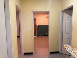 Um corredor com uma porta à direita, um quarto visível no final e um tábua de passar roupa colorida na parede da direita.