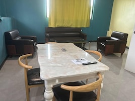 Uma sala de reuniões com uma mesa de mármore, quatro cadeiras de madeira, um sofá e uma cortina amarela.
