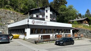 A modern hotel with a sign reading "Auberge de l'Ours." It features a two-story structure with a flat roof, large windows, and a stone retaining wall. - Auberge de l'Ours (Vex)