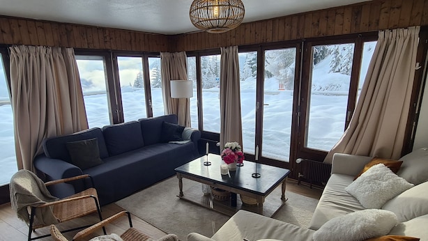 A cozy room with a dark blue sofa, a white sofa, wicker chairs, and a wooden coffee table. Large windows offer a view of a snowy landscape. - Auberge de l'Ours (Vex)