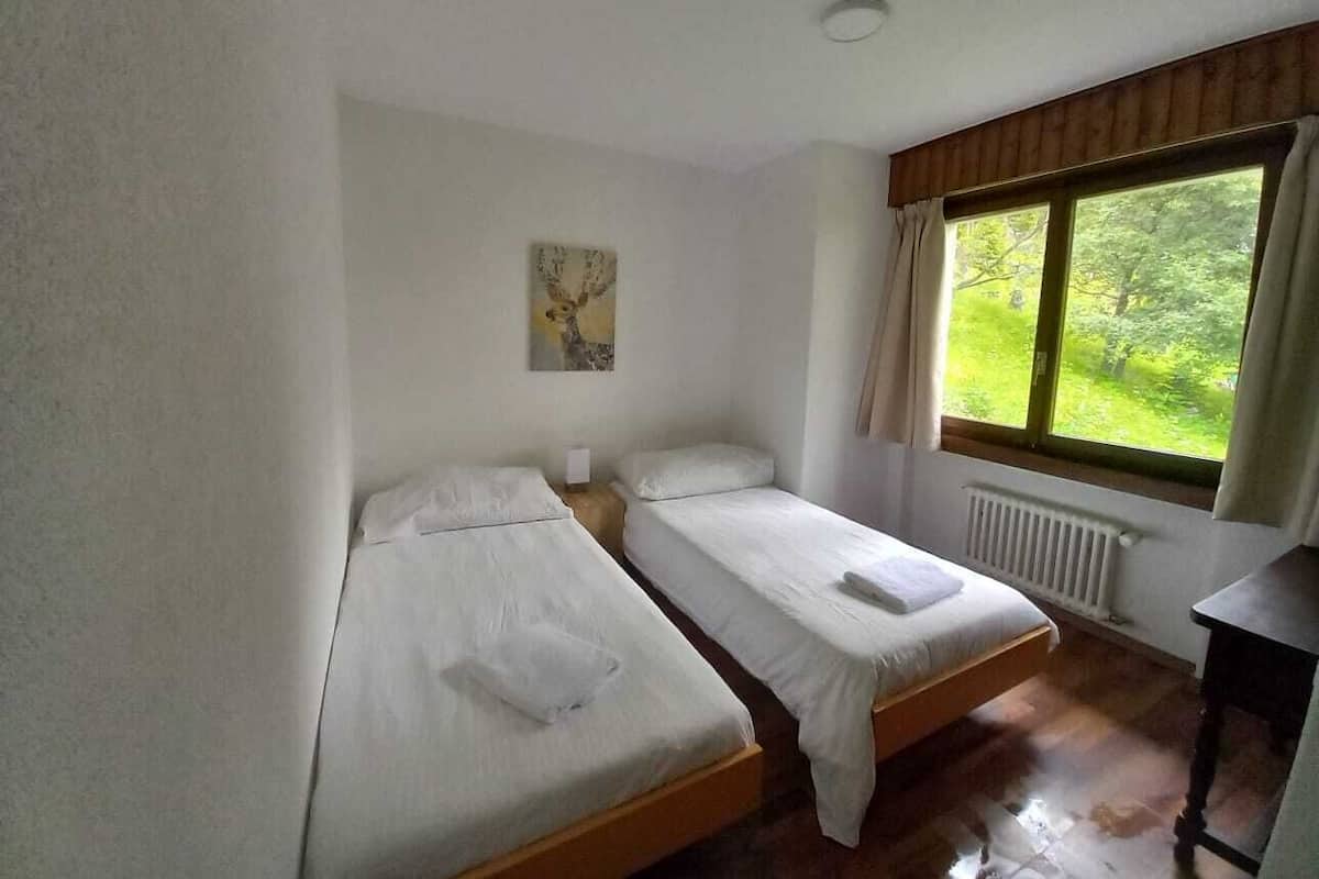 A room with two single beds, a small table, a radiator, and a window with a view of greenery.