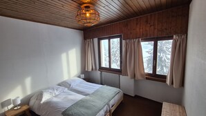 A bedroom with a wooden bed, a bedside table with a lamp, a window with curtains, and a view of snow-covered trees. - Auberge de l'Ours (Vex)