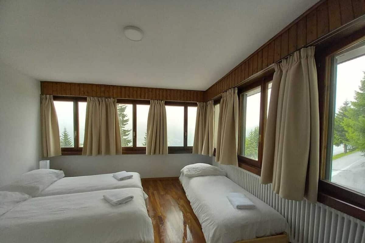 A room with two single beds, a wooden floor, large windows with curtains, and a radiator.