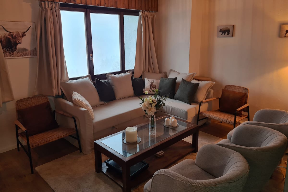 A cozy living room with a sofa, armchairs, a coffee table, and a large window with curtains.