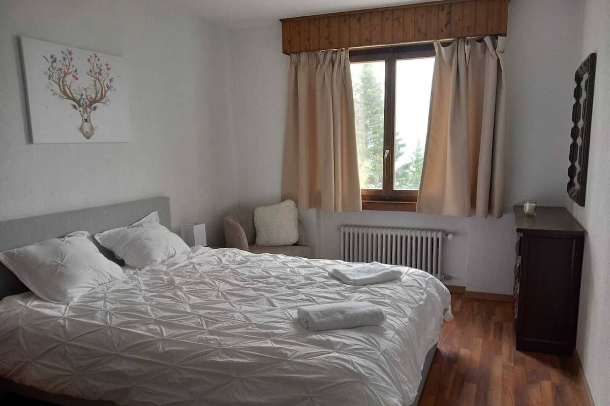 A bedroom with a large bed, a window with curtains, a radiator, and a small cabinet.