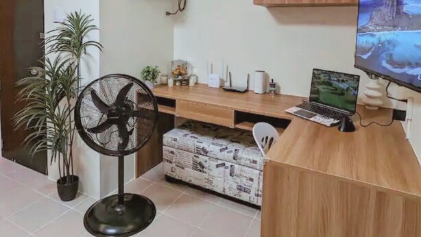Ein moderner Arbeitsplatz mit Schreibtisch aus Holz, Computer und Ventilator. An der Wand befindet sich eine weiße Klimaanlage und ein Blumentopf.