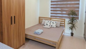 Ein Schlafzimmer mit Bett, Holzschrank, Fenster mit Jalousien, Deckenleuchte und einer Zimmerpflanze.