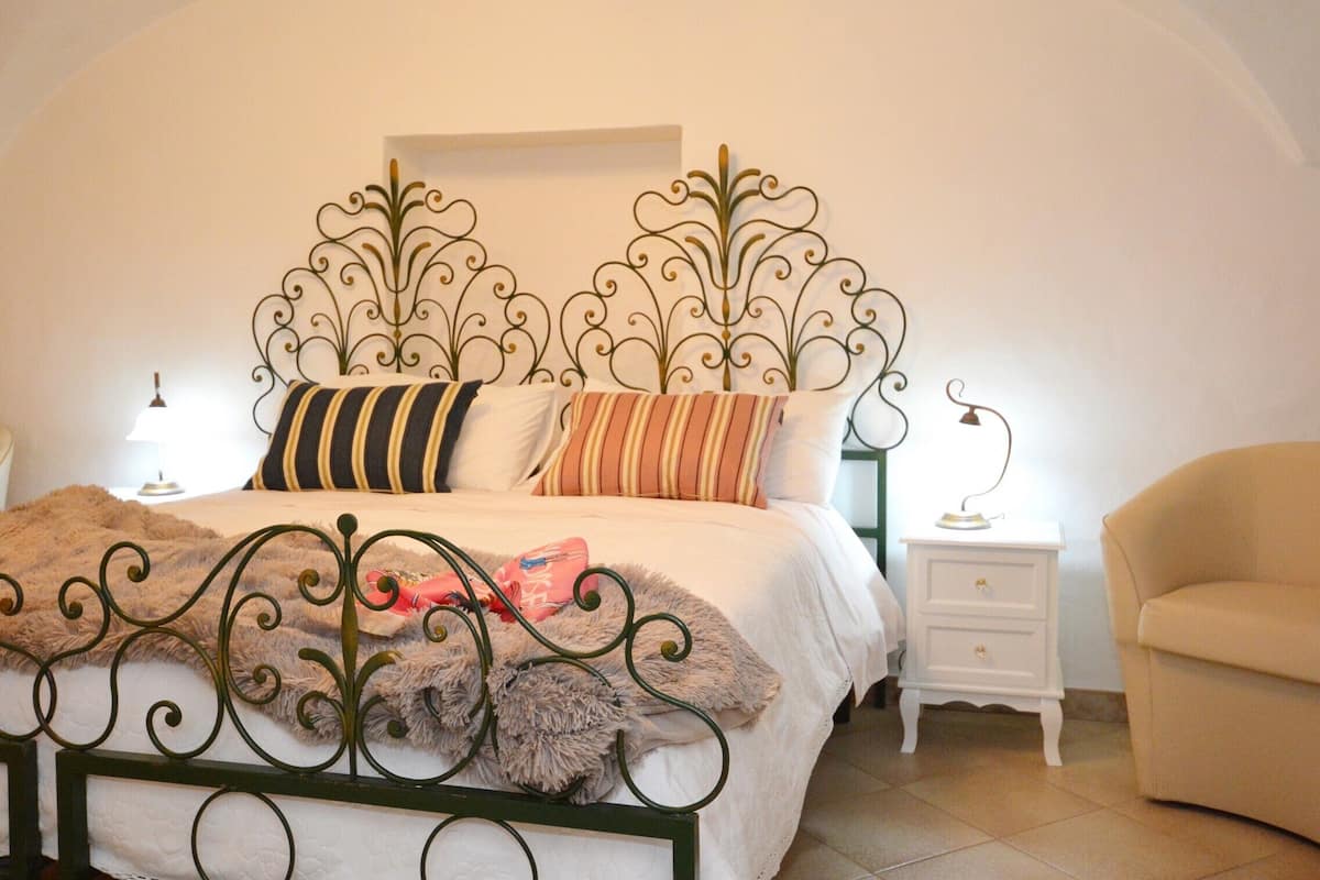 A neatly arranged bedroom with a metal bed frame, a white bedside table with a lamp, and a beige armchair.