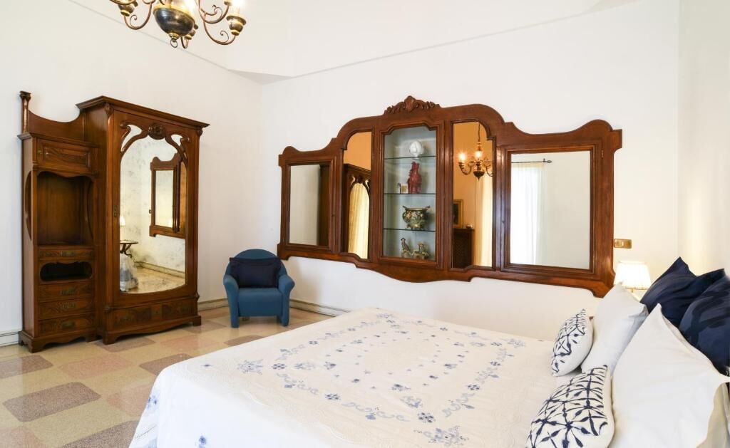 A bedroom with a large bed, a wooden wardrobe, a chair, and a mirror with shelves.