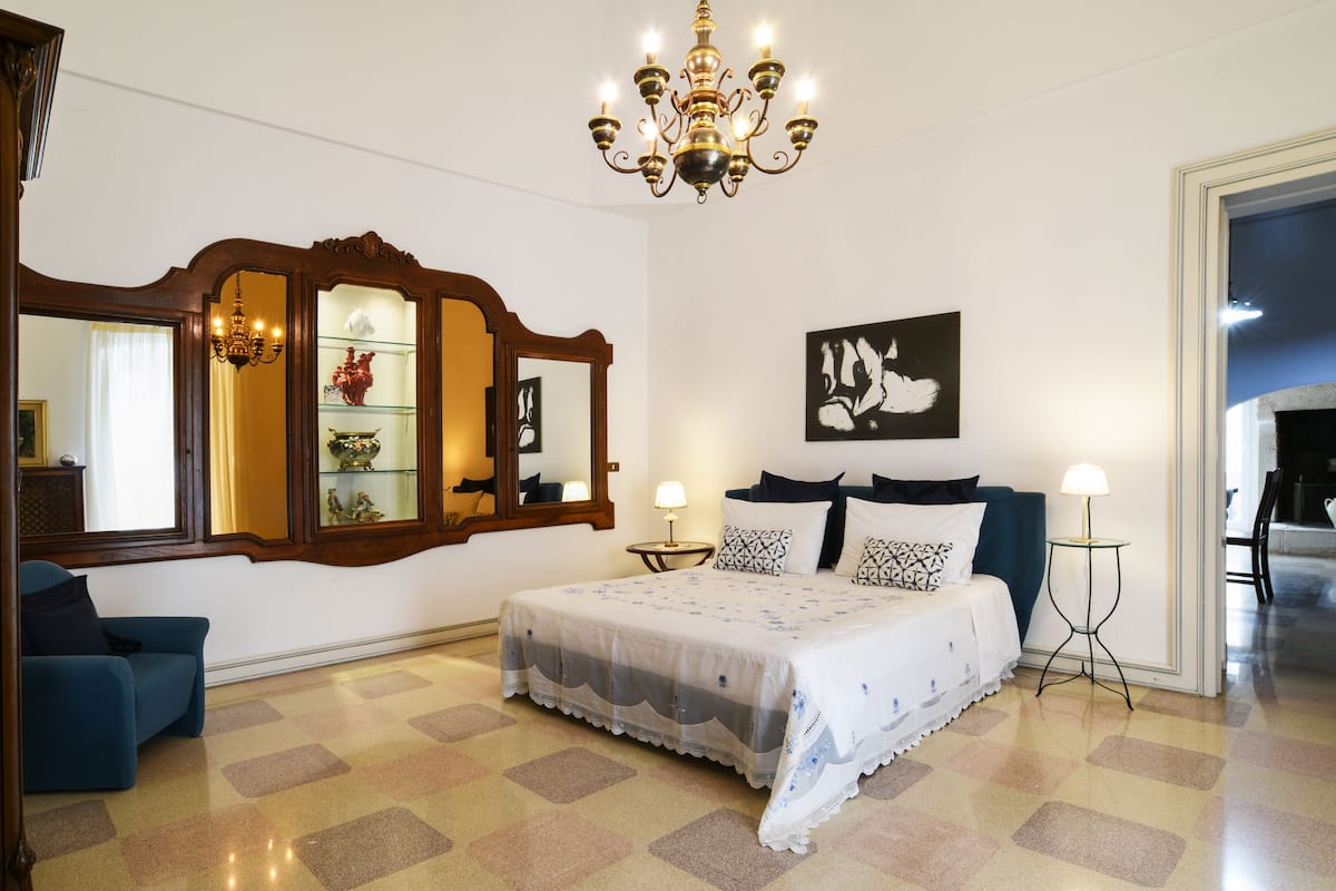 A bedroom with a bed, a dresser, a mirror, a chandelier, and a blue armchair.