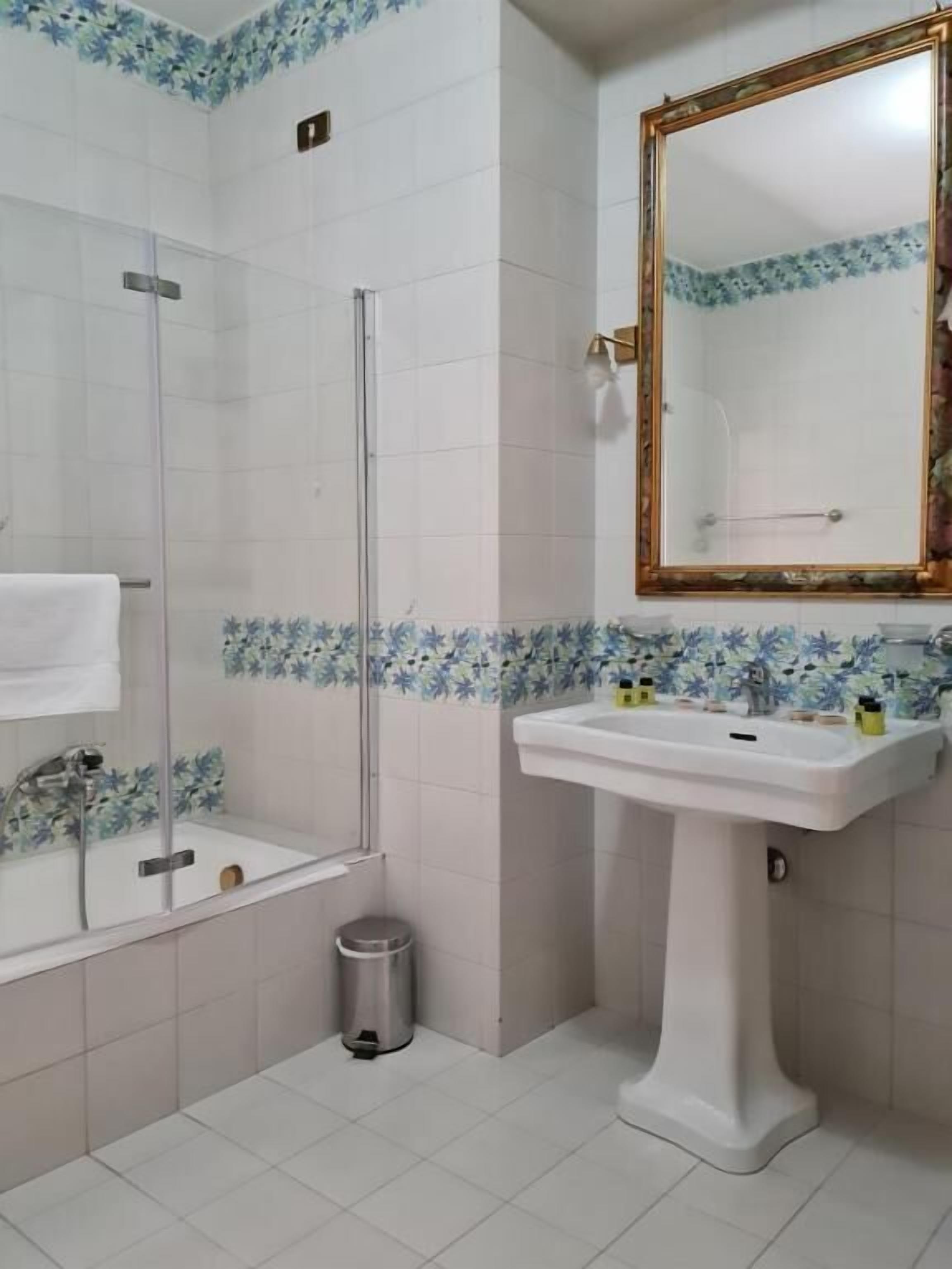 A bathroom with a glass-enclosed shower, a pedestal sink, and a mirror with a gold frame.