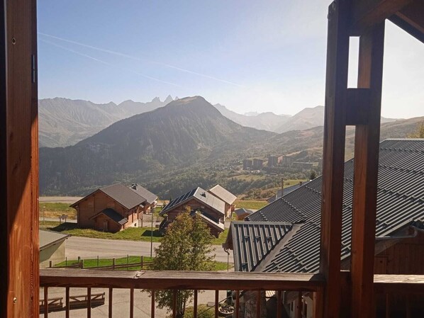 Vista desde un balcón sobre un paisaje montañoso con varias construcciones, incluyendo una cabaña de madera y una casa moderna con paneles solares.