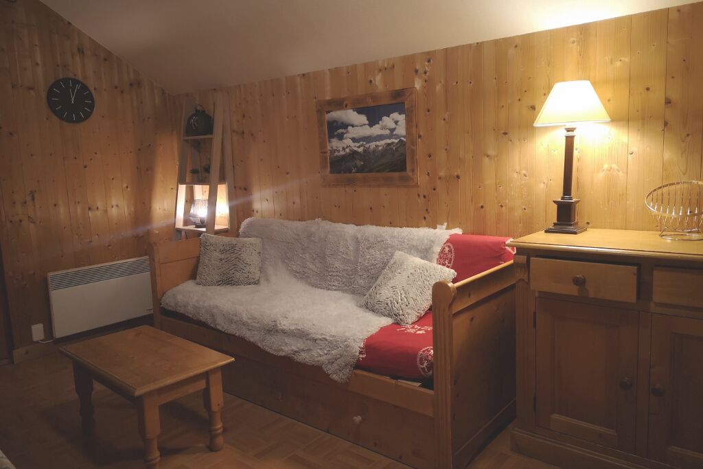 Une pièce chaleureuse avec un plancher en bois, un canapé recouvert d’une couverture blanche, une petite table en bois, une lampe et une horloge au mur.