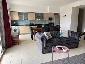 A modern living room with a grey sofa, a small pink table, and a dining area with a wooden table and chairs.