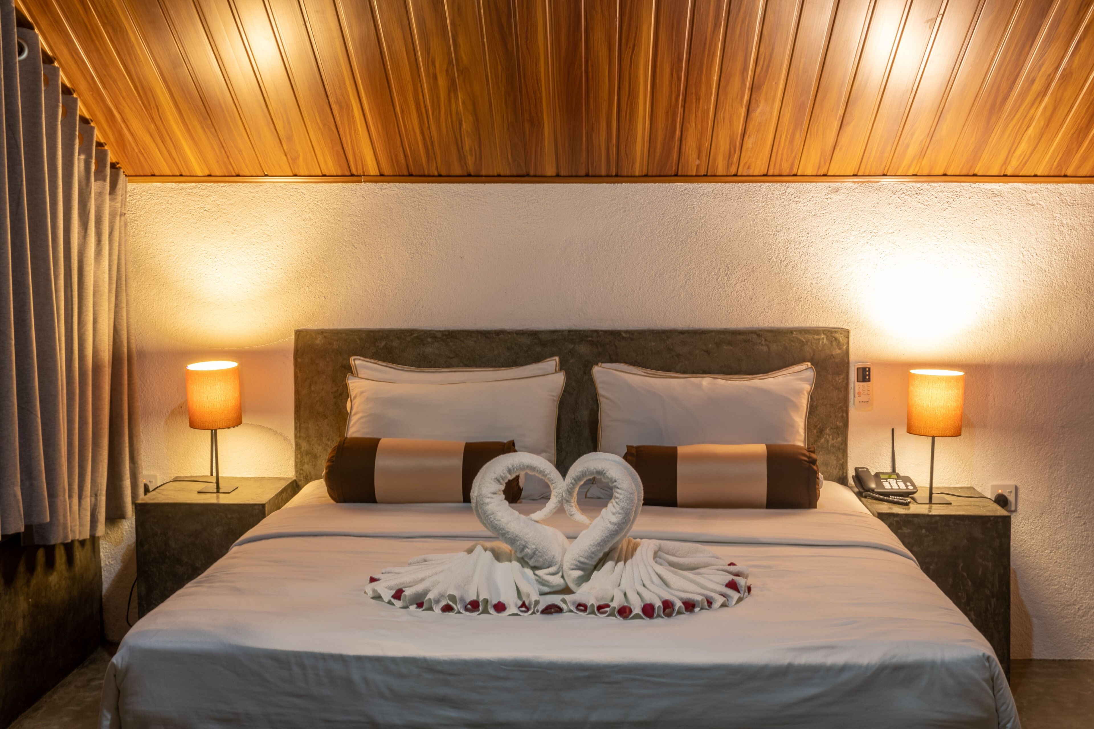A hotel room with a neatly made bed, two bedside lamps, and a heart-shaped towel arrangement on the bed.