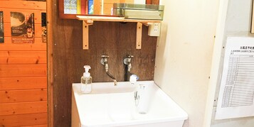Un baño con un lavamanos blanco, dos llaves, un dispensador de jabón y un estante de madera sobre el lavamanos.