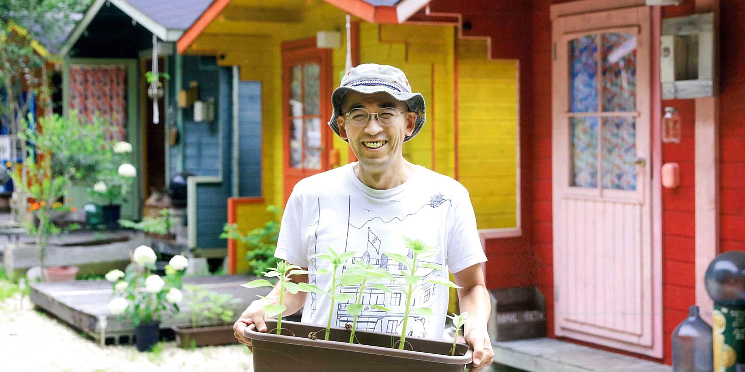 A person holding a tray with potted plants in front of a colorful building with a red door and a window with a patterned curtain.