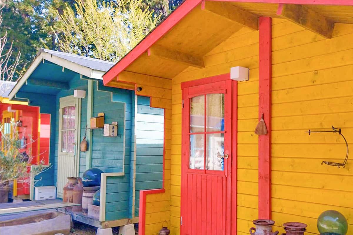 Une petite cabane en bois aux couleurs vives, avec une porte rouge et une porte bleue, entourée de divers objets, comme un balai, une lanterne et un verre vert.