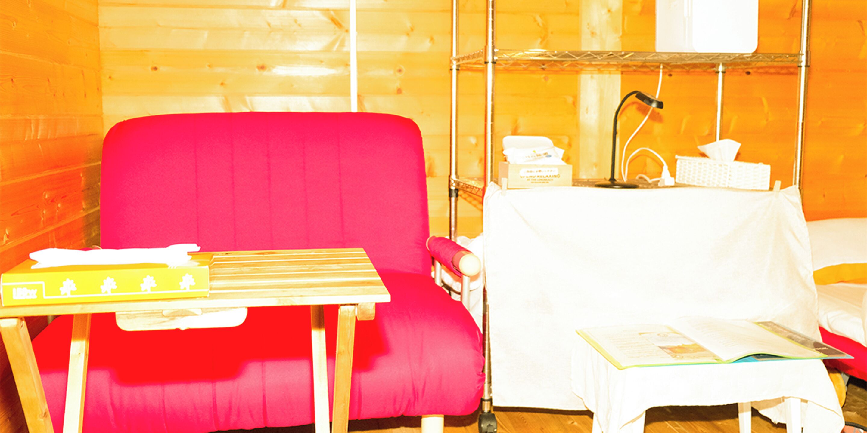 Una habitación con una silla roja, una mesita pequeña con un libro y una mesita de noche blanca con una lámpara y pañuelos.