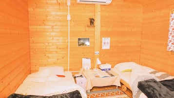 A small, cozy room with wooden walls and two beds. There is a small table with a lamp, a heater, and a few personal items.