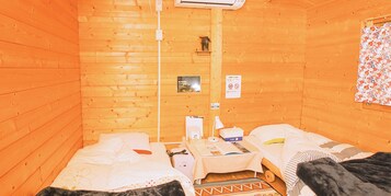 Una habitación pequeña y acogedora con paredes de madera y dos camas. Hay una mesita con una lámpara, un calentador y algunos objetos personales.