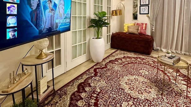 A living room with a large flat-screen TV displaying a Netflix interface, a red patterned rug, a potted plant, and a brown sofa. - EXTACODE APARTMENT (Abuja)