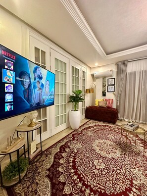 A living room with a large flat-screen TV displaying a Netflix interface, a red patterned rug, a potted plant, and a brown sofa. - EXTACODE APARTMENT (Abuja)