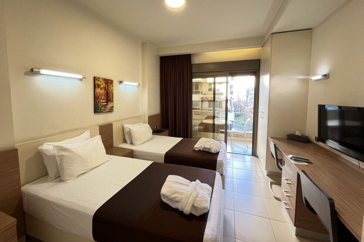 A hotel room with two beds, a desk with a television, and a view of the outdoors through a glass door.