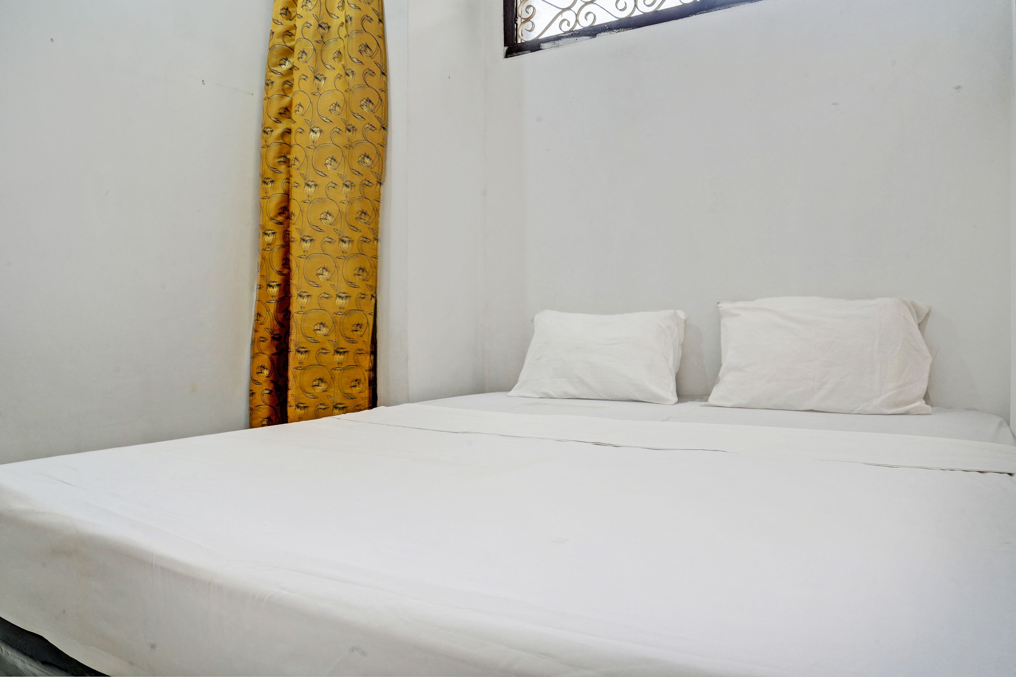 A single bed with white bedding in a room with plain white walls and a yellow curtain with a floral pattern.