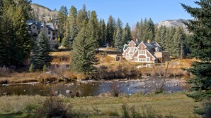 A large house with a river in front, surrounded by trees and a mountain in the background.