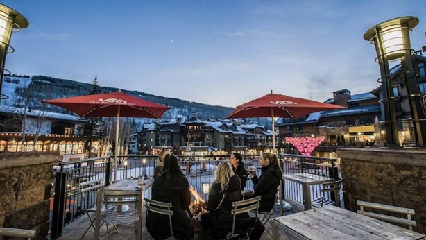 An outdoor seating area with tables and chairs, red umbrellas, and a view of a snowy mountain landscape.