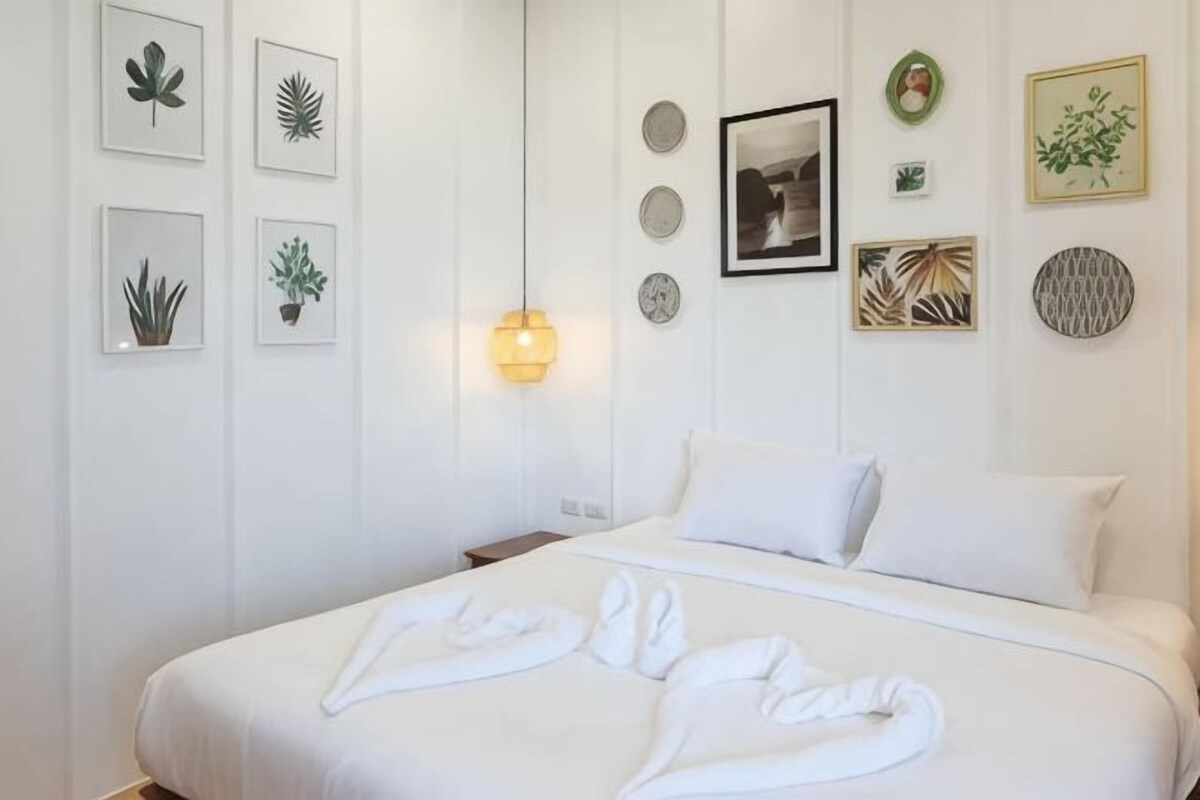 Habitación de hotel con cama, ropa de cama blanca, una lámpara de pared e impresiones botánicas enmarcadas.