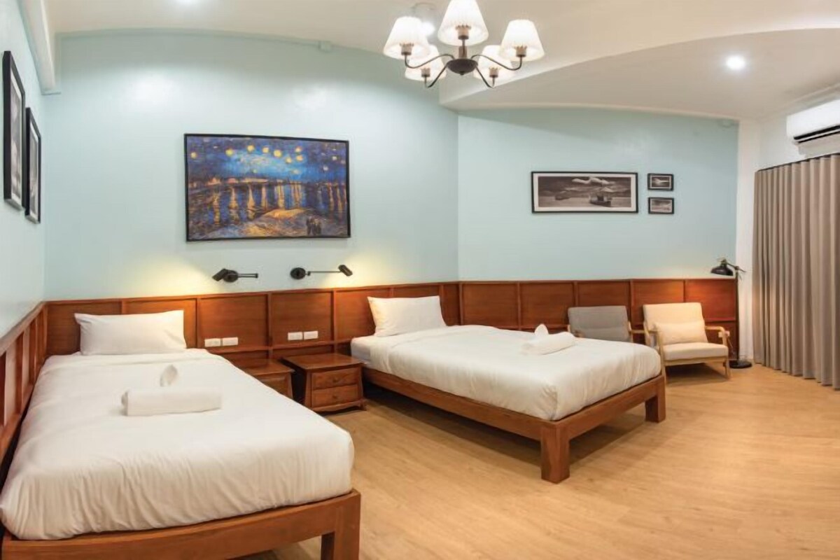 Habitación de hotel con dos camas, cabeceros de madera, una mesita de noche, una silla y un cuadro en la pared.
