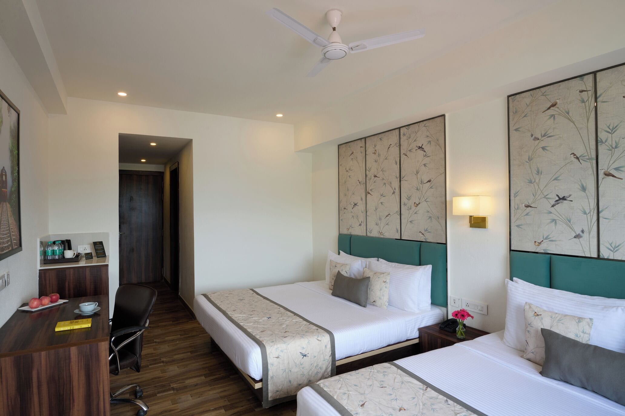 A hotel room with two beds, a desk, a chair, a ceiling fan, and artwork on the walls.