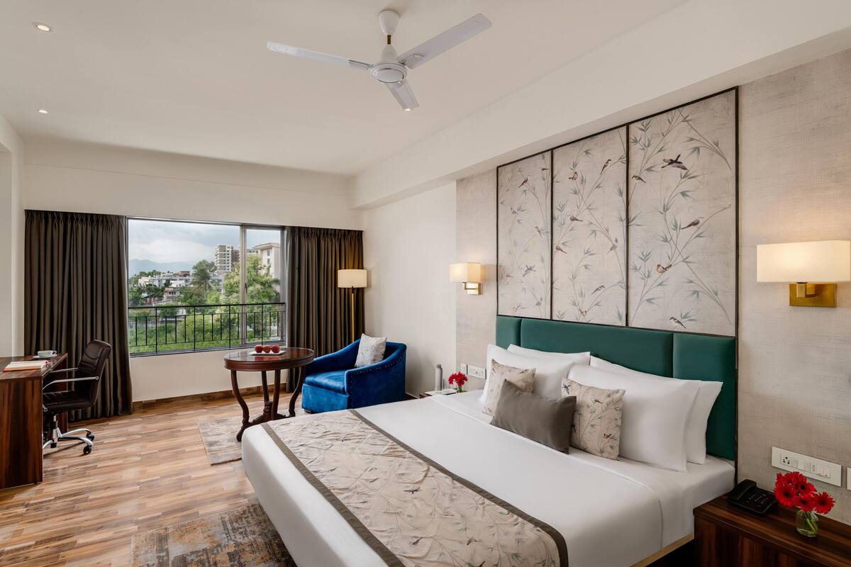 A hotel room with a large bed, a desk with a chair, a small table, and a blue armchair. There is a ceiling fan, a window with curtains, and a view of buildings outside.