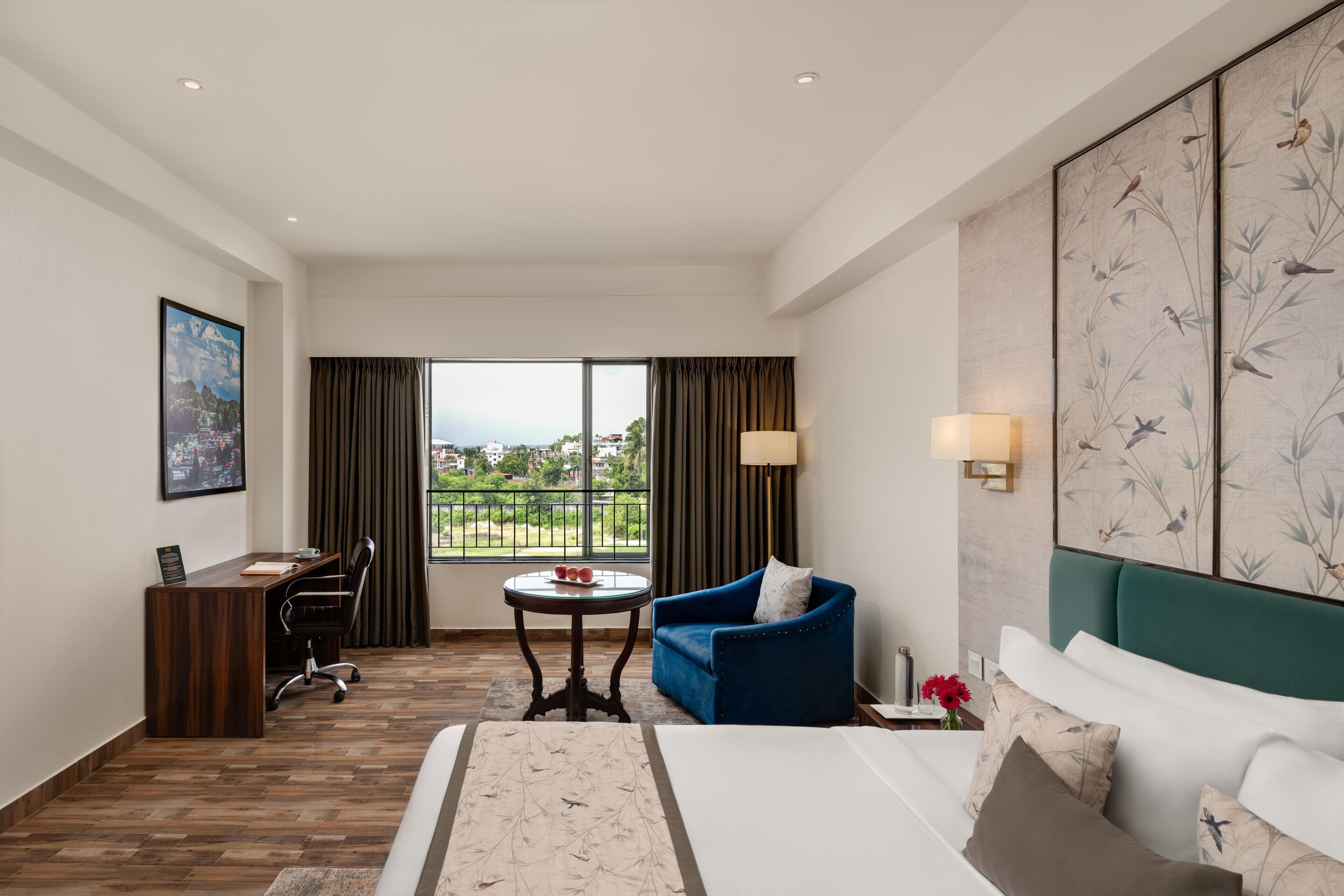 A hotel room with a bed, a desk, a chair, a small table, a sofa, a view of the outdoors, and a large artwork on the wall.