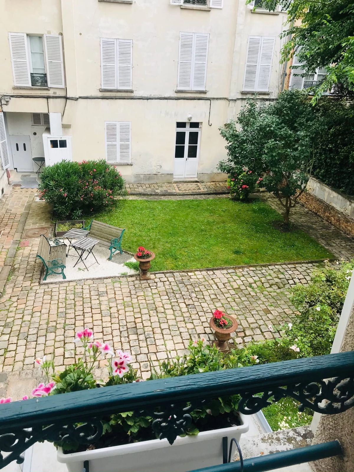 Une cour avec une terrasse dallée, une petite pelouse et un bâtiment aux volets blancs.