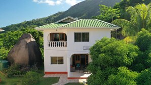 Une maison à deux étages, dotée d’un toit végétalisé, entourée d’une végétation luxuriante et d’une grande formation rocheuse.