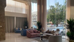 A spacious lobby with a blue sofa, a pink sofa, and a grey armchair. There are large windows with curtains, a chandelier, and a potted plant. - SAVOYE HOTEL (Jeddah)