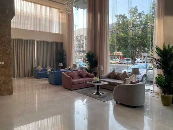 Un hall espacioso con un sofá azul, un sofá rosa y un sillón gris. Se observan amplias ventanas con cortinas, un candelabro y una planta en maceta.