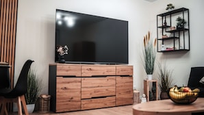 Un salon moderne avec un téléviseur à écran plat posé sur un meuble en bois, un fauteuil noir, une petite table sur laquelle se trouve un panier de fruits, et une étagère décorative agrémentée de plantes et de livres.