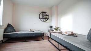 Une chambre à coucher au style minimaliste, avec deux lits, une petite table de chevet, une étagère fixée au mur et un radiateur.