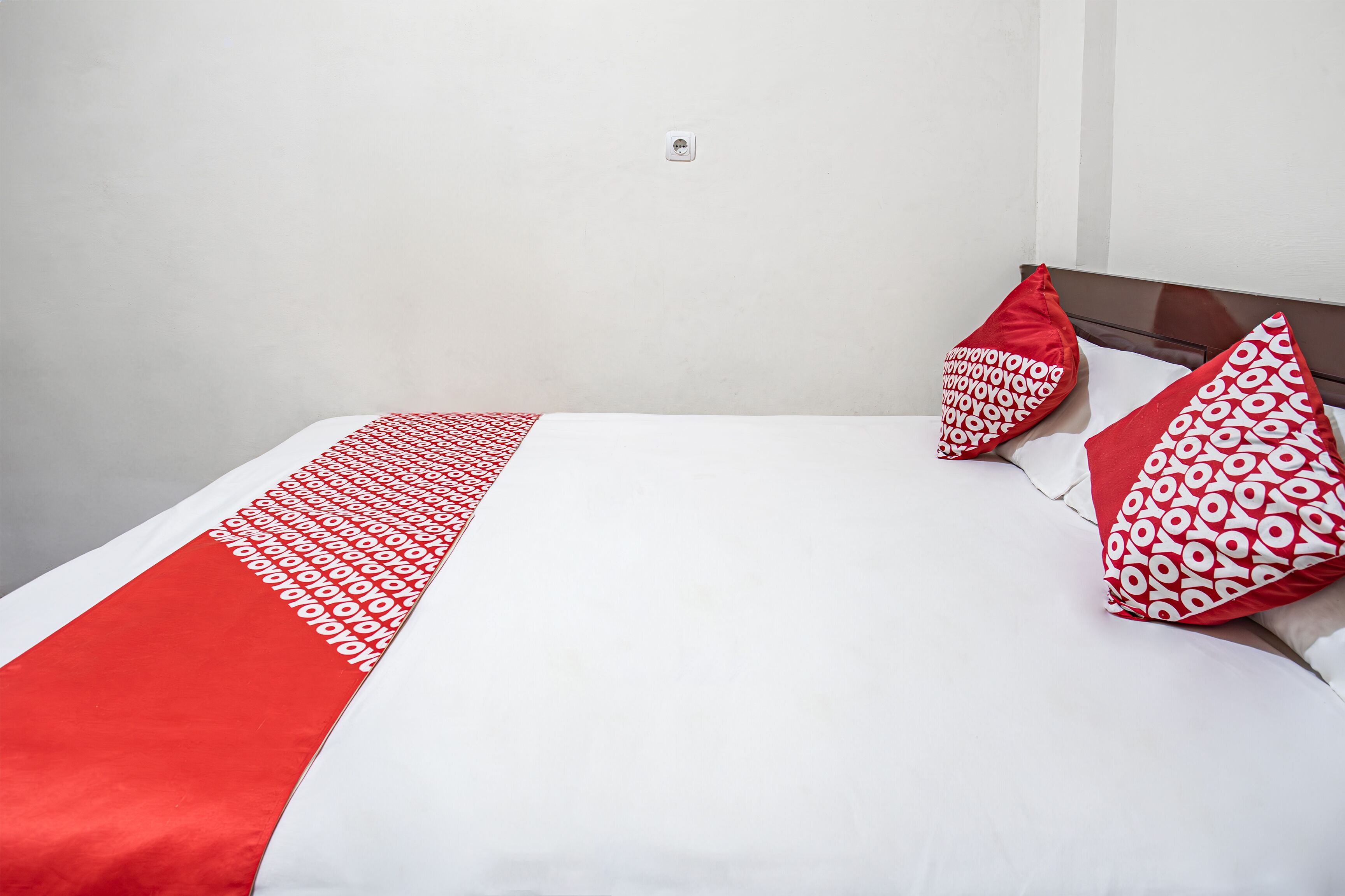 A bed with white and red bedding, two red pillows with a circular pattern, and a white wall with a power outlet.