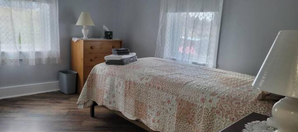 A bedroom with a bed, a dresser, a lamp, and a window with sheer curtains.