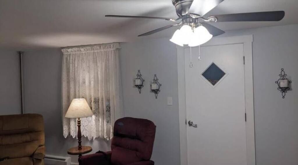 A room with a ceiling fan, a window with lace curtains, a lamp, a chair, and wall-mounted sconces.