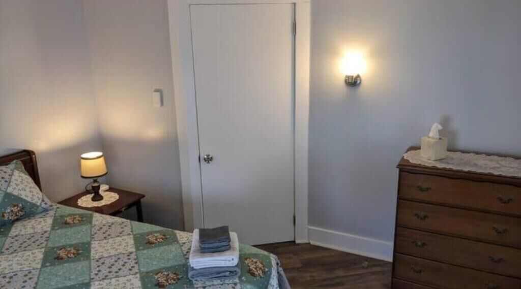 A bedroom with a bed, a nightstand with a lamp, a dresser, and a door.