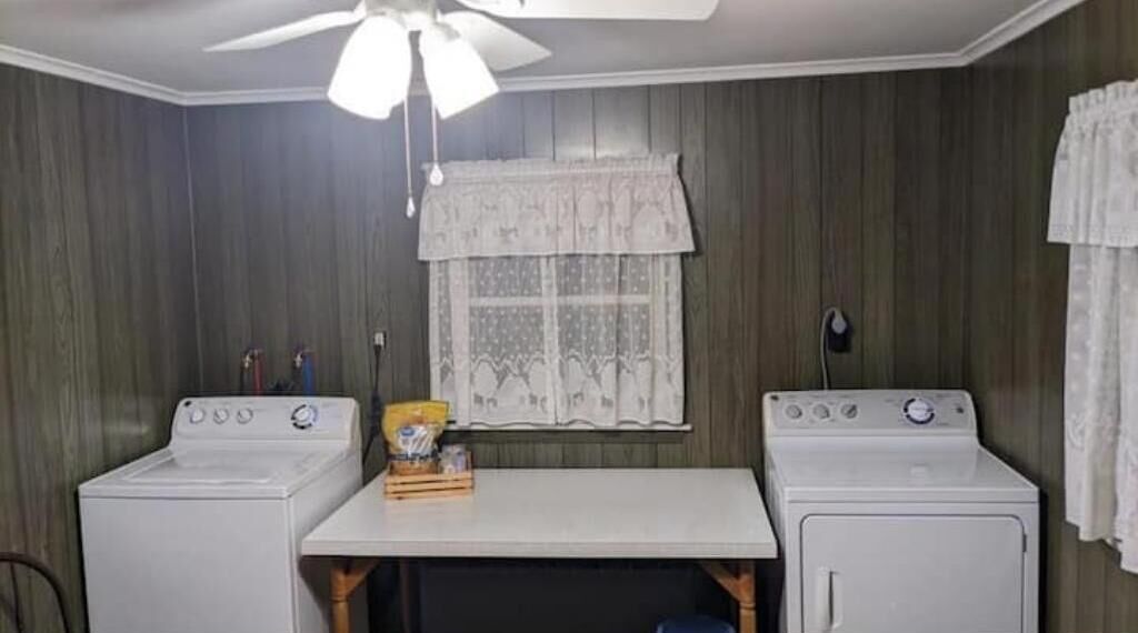 A laundry room with two white washing machines, a ceiling fan, and a window with a lace curtain.