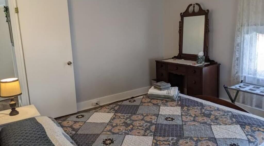 A hotel room with a bed, a bedside table with a lamp, a dresser with a mirror, and a patterned floor.